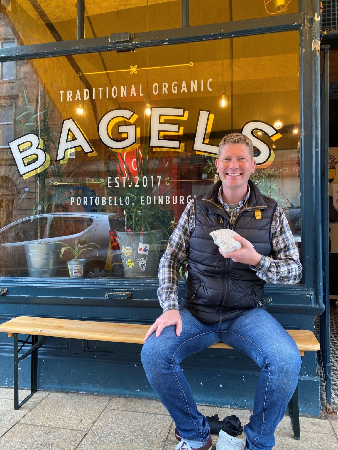 A man sits smiling on a bench in front of a bagel shop window in Edinburgh, holding a wrapped bagel. The shop sign reads Traditional Organic Bagels.