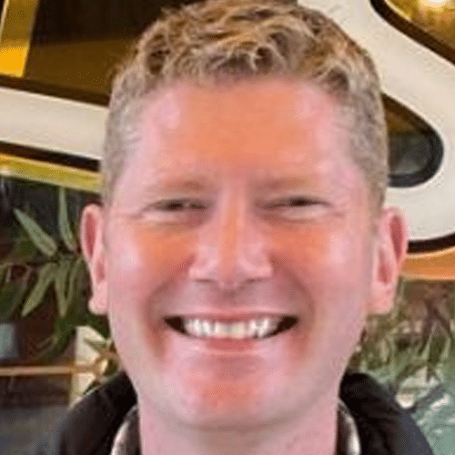A person with short, curly blond hair is smiling broadly. Green leafy plants and part of a large sign with the letter S are visible in the background.
