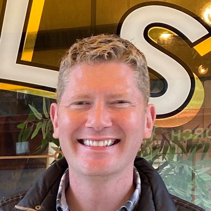 A man with short, curly blond hair smiles broadly in front of a sign with large white and yellow letters. Green leaves are visible in the background.