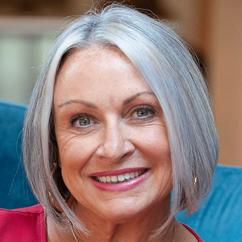 Smiling older woman with short, straight, grey hair and blue eyes, wearing a pink top. She is sitting indoors in front of a blue background.
