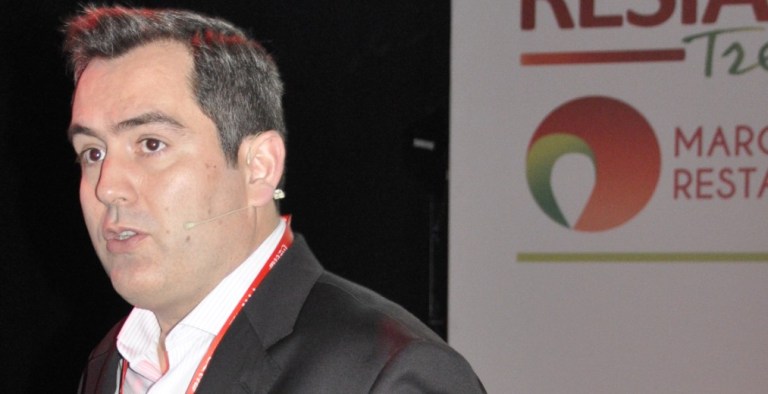 A man in a suit speaks on stage with a microphone headset. Behind him is a partial sign with a colourful logo and partial text.