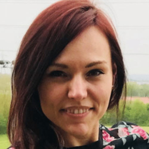 A woman with long brown hair smiles at the camera outdoors, with greenery and a cloudy sky in the background. She is wearing a floral-patterned garment.