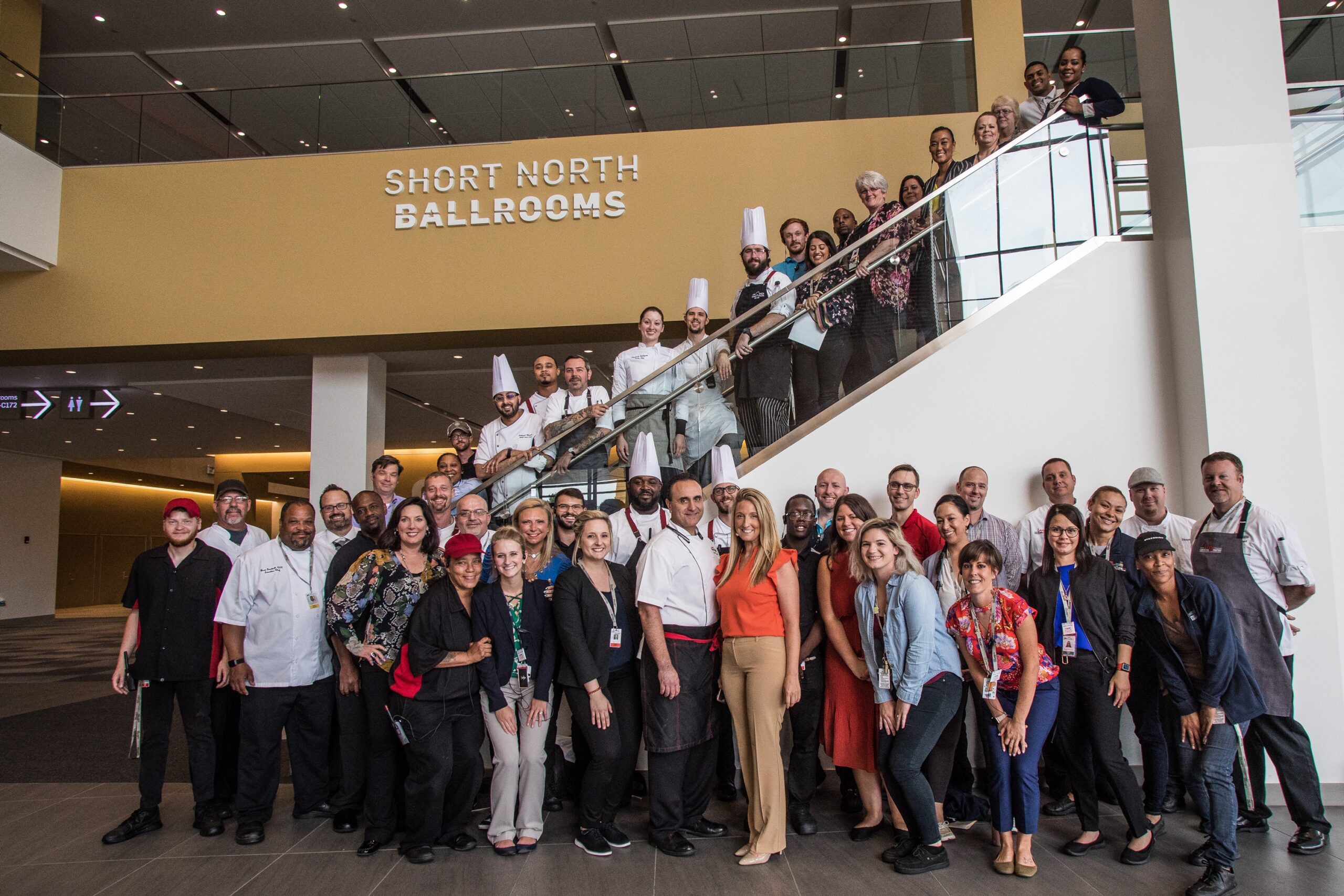A large group of people, including chefs in uniforms, pose together on a staircase and floor under a sign reading Short North Ballrooms inside a modern building.