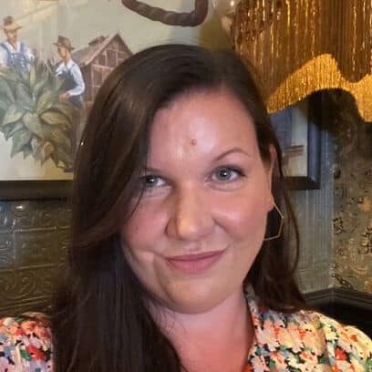 A woman with long brown hair, wearing a floral top, smiles at the camera indoors. Behind her is a painting of two people and part of a fringed lampshade.