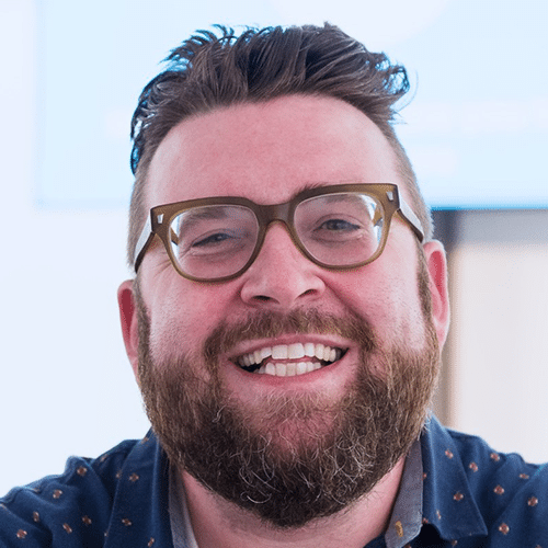 A bearded man with glasses smiles warmly at the camera, wearing a dark blue shirt with a blurred light background.