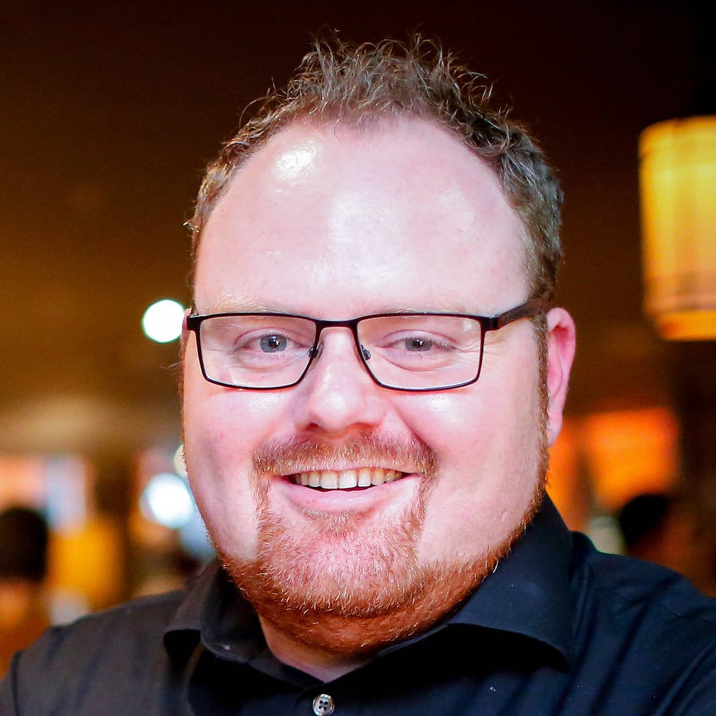 A man with short curly hair, glasses, and a red beard smiles at the camera. He is wearing a black shirt and standing indoors with warm lighting in the background.