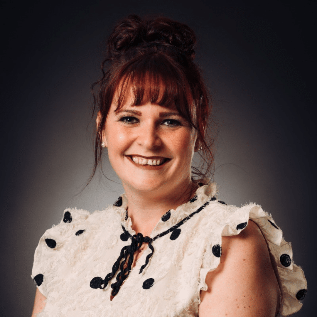 A woman with auburn hair in an updo, wearing a white ruffled top with black polka dots, smiles in front of a dark gradient background.