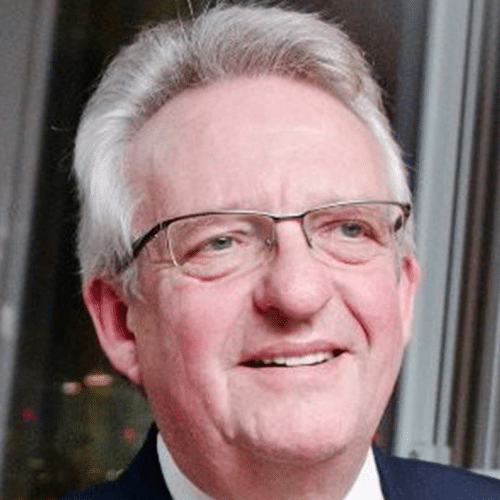 A smiling older man with grey hair and glasses, wearing a suit, stands in front of a window with city lights blurred in the background.