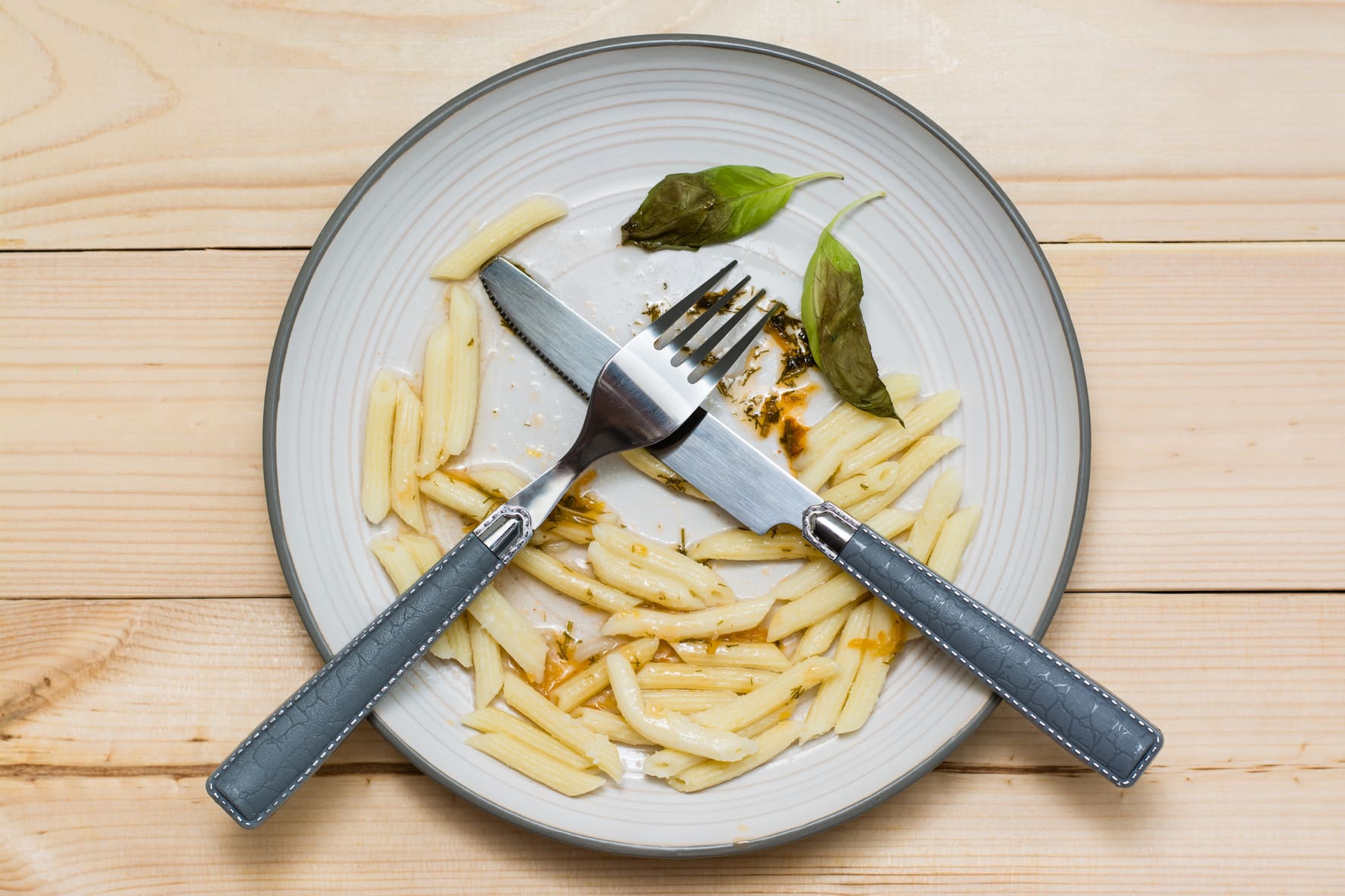 A plate with leftover penne pasta, sauce stains, and two basil leaves. A fork and knife are crossed on the plate, which sits on a light wooden table.
