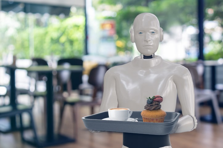 A humanoid robot holds a tray with a cup of coffee and a chocolate cupcake topped with cream and a strawberry in a modern, brightly lit café.