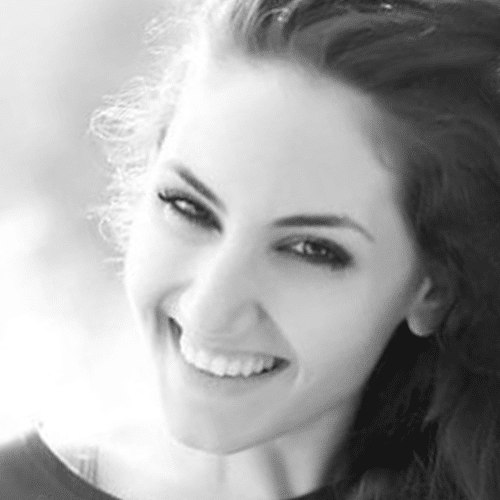 A young woman with long dark hair smiles brightly at the camera. The image is black and white, with a soft, blurred background.