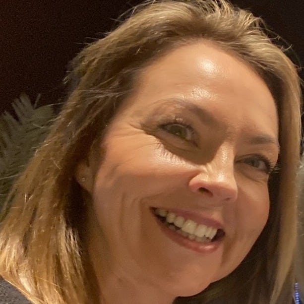 A woman with light brown hair smiles brightly at the camera against a dark background.