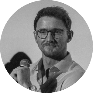 A man with short hair, glasses, and a beard is holding a microphone and wearing a buttoned shirt. The image is black and white and framed in a circular crop.