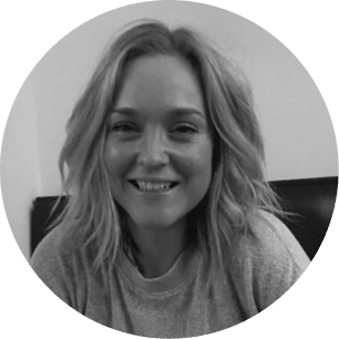 A woman with shoulder-length wavy hair smiles at the camera whilst sitting indoors. The image is in black and white and framed in a circular border.
