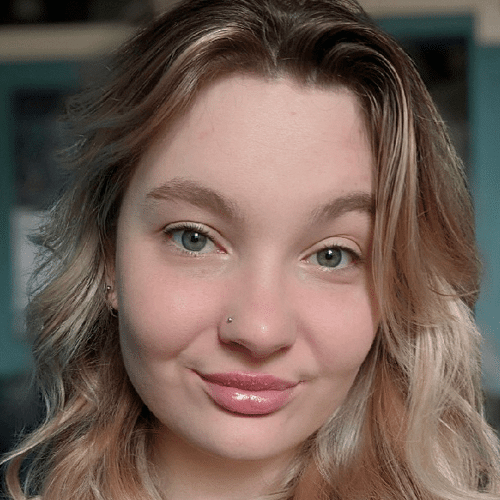 A young woman with wavy blonde hair and light eyes smiles softly at the camera. She has a nose piercing and is in a softly lit indoor setting with a blue background.