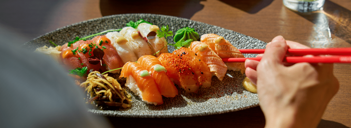 A person uses red chopsticks to pick up a piece of assorted sushi from a grey plate, which also has ginger and wasabi, on a wooden table in bright natural light.