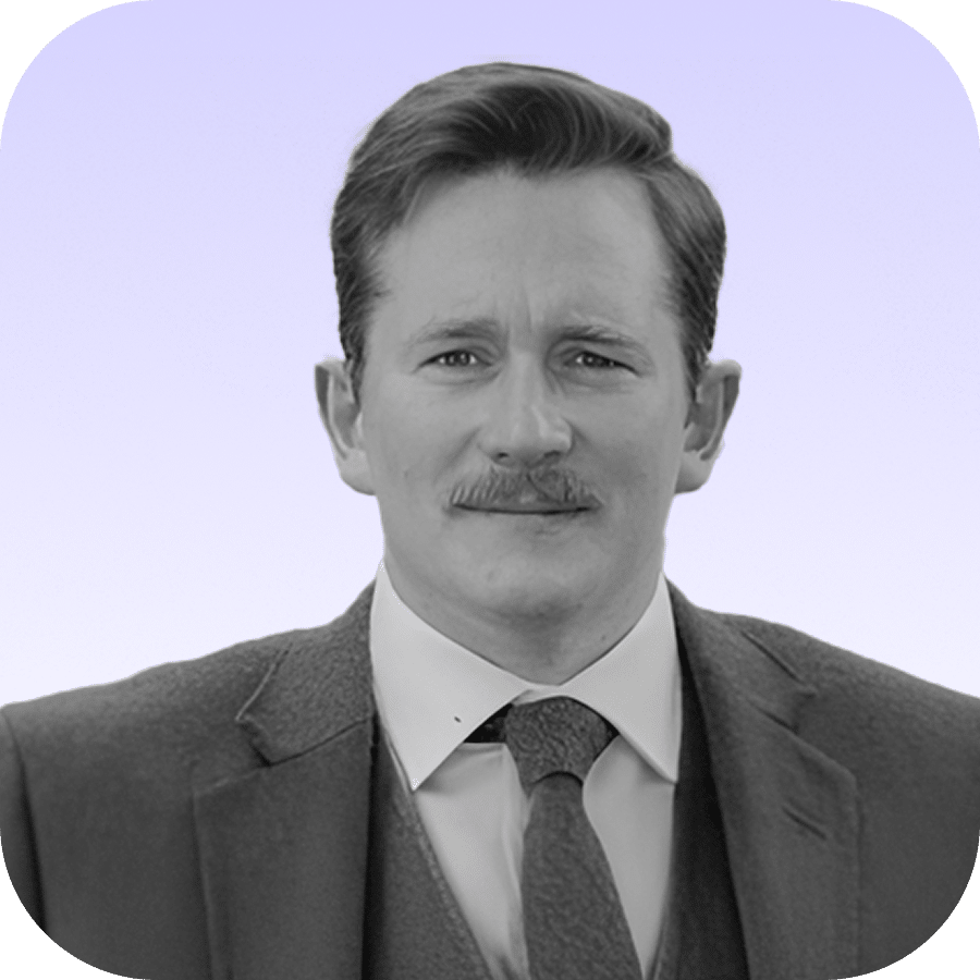 A man in a suit and tie with short hair and a moustache, looking at the camera, against a light purple gradient background. The image is in black and white with rounded corners.