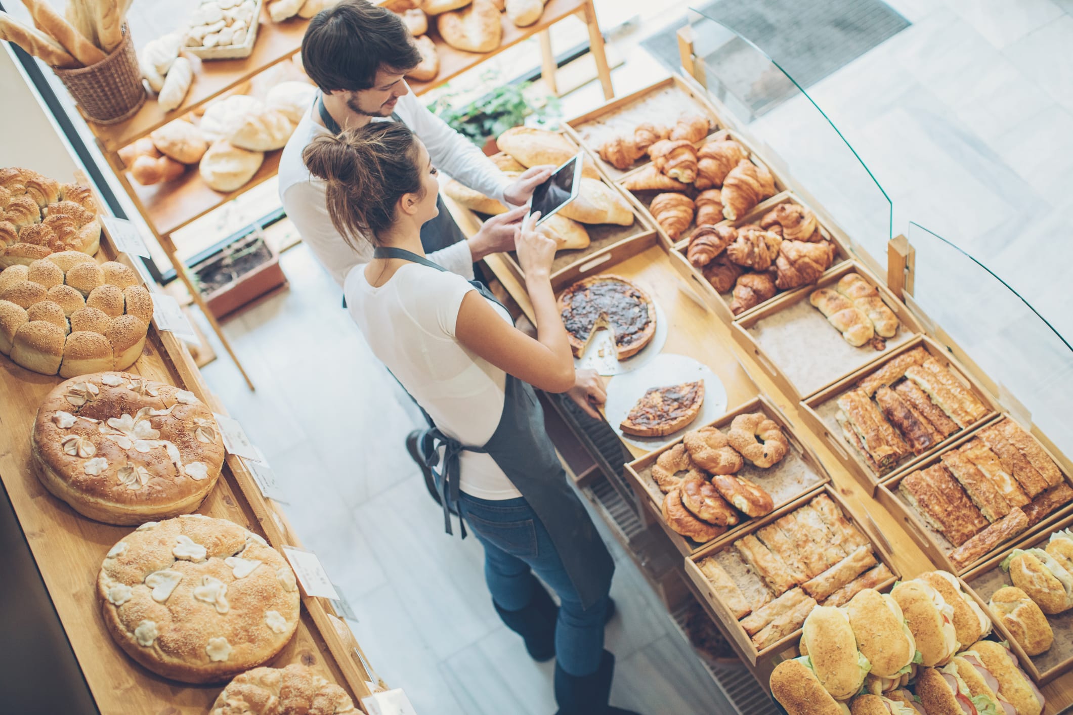 Two people in a bakery review an order on a tablet whilst surrounded by shelves full of fresh bread, pastries, and baked goods.