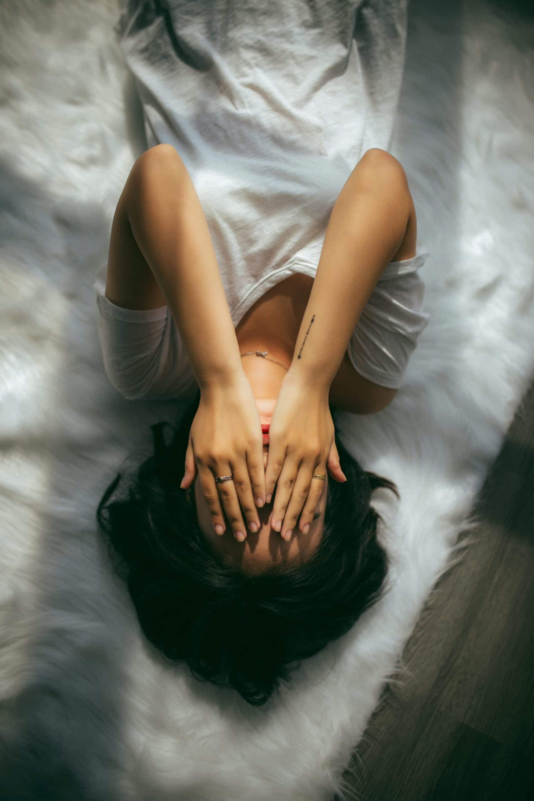 A person in a white shirt lies on a fluffy white rug with both hands covering their face, as sunlight filters through, casting soft shadows.