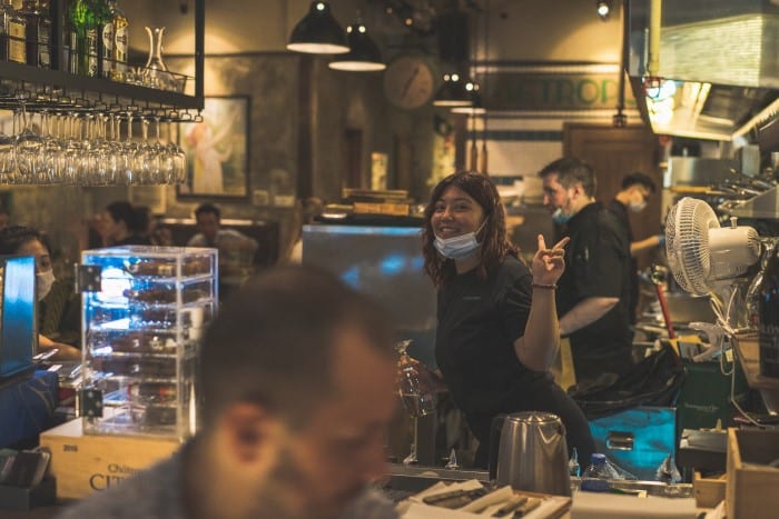 A woman wearing a mask around her chin flashes a peace sign in a busy restaurant or bar, with other staff and customers in the background.