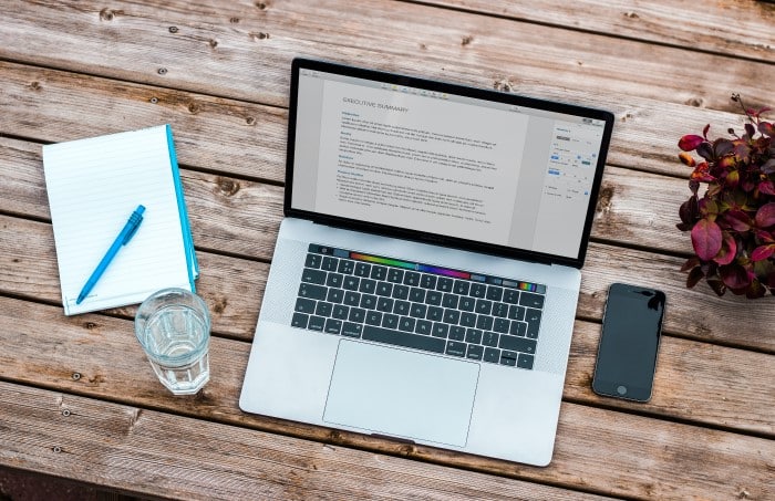 A laptop displaying a document sits on a wooden table with a notebook, pen, glass of water, mobile phone, and a plant nearby.