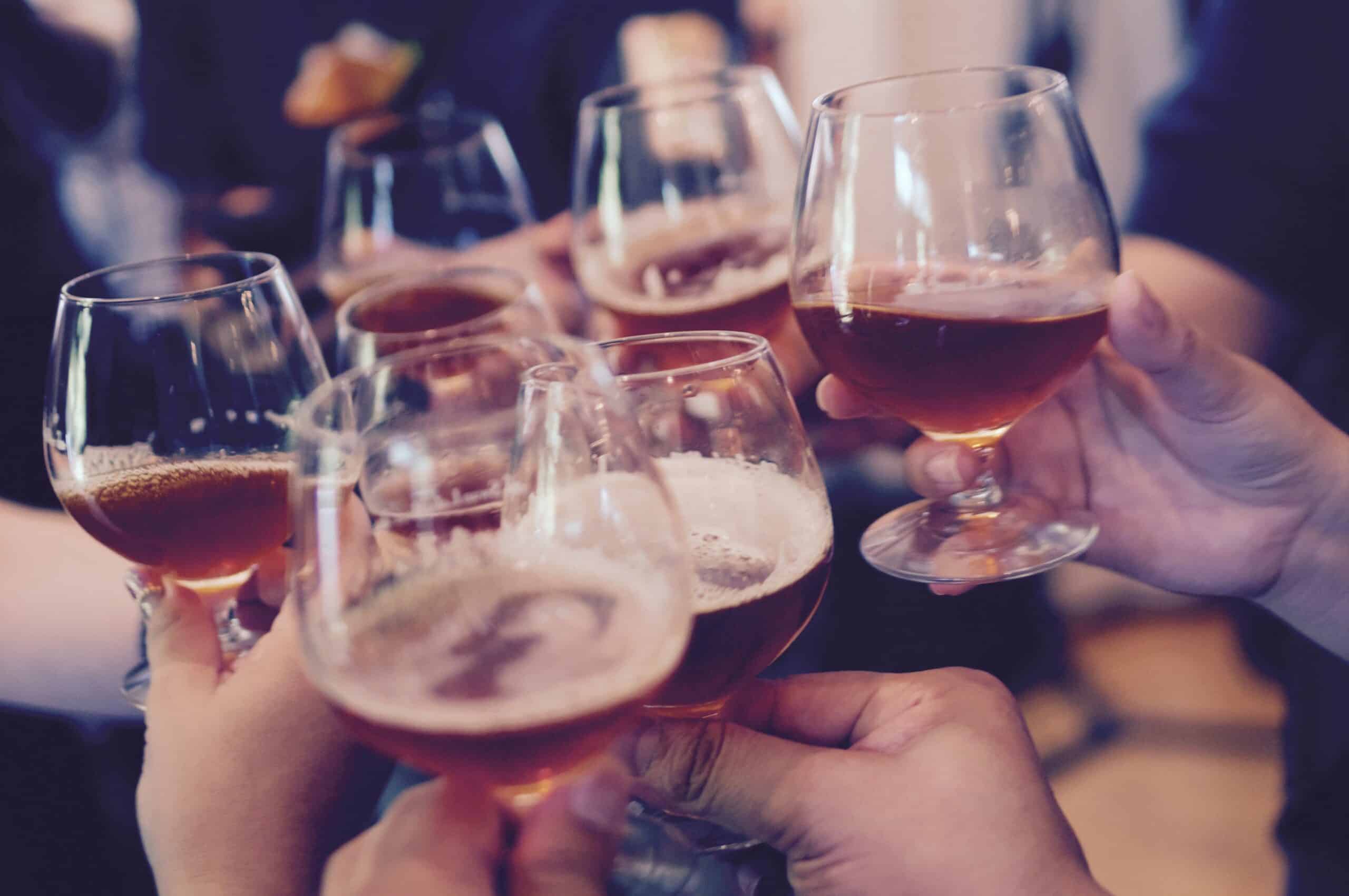 Several hands holding snifter glasses filled with amber-coloured beer or liquor, clinking them together in a celebratory toast, with a blurred background.